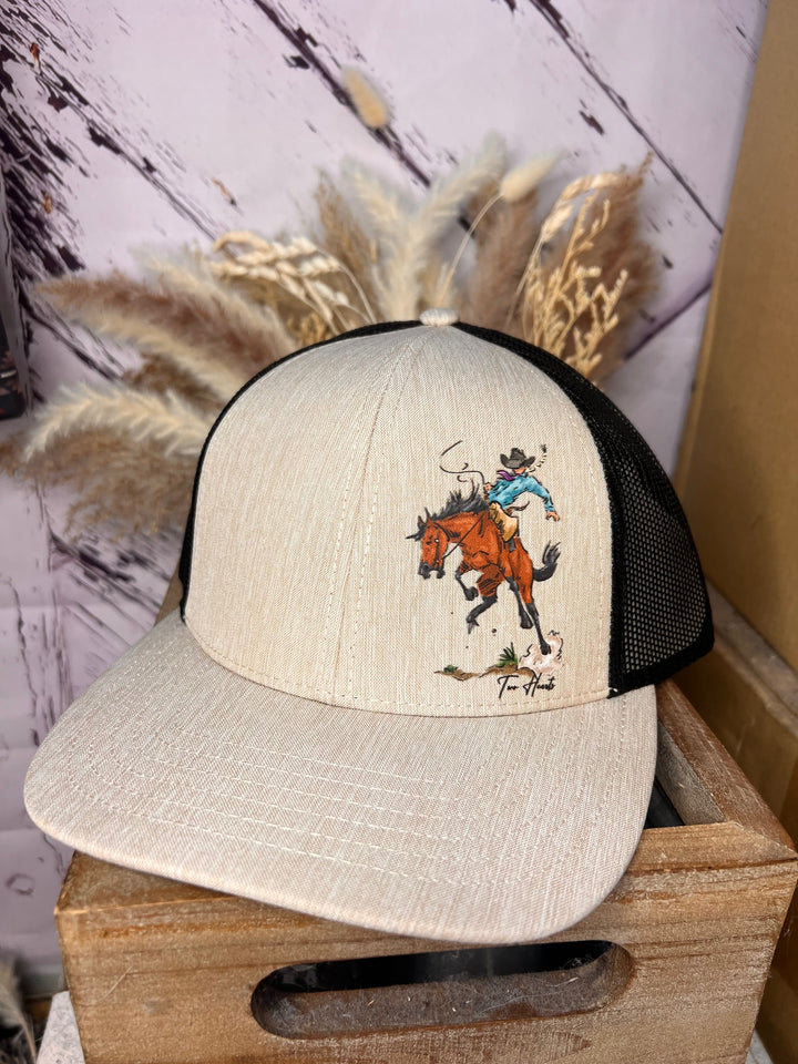 A beige and black trucker hat with a hand-drawn design of a cowgirl riding a bronc horse, held by a person against a wooden backdrop.