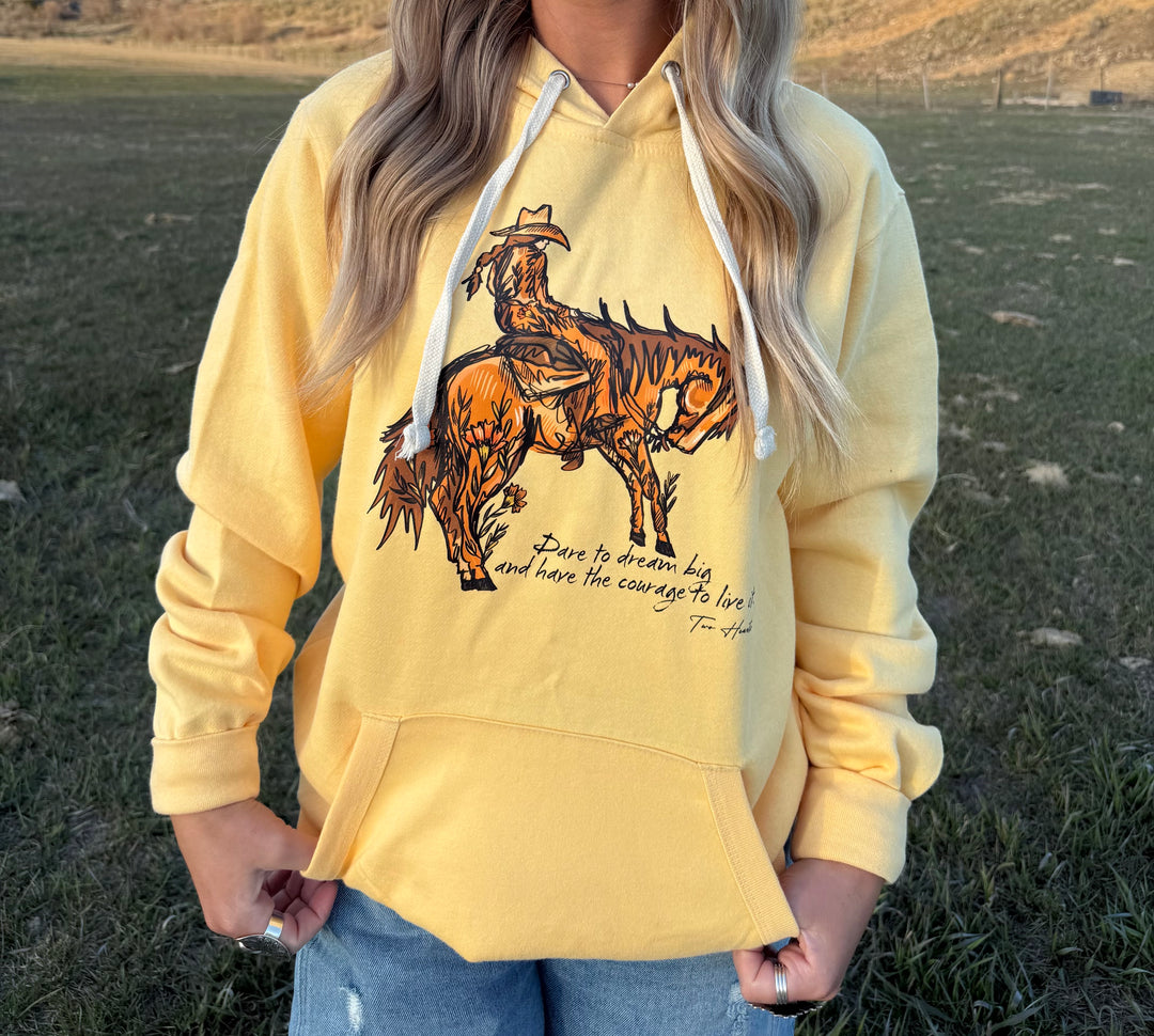 A person wearing a yellow hoodie with a graphic of a cowgirl riding a bucking horse and the text 'Dare to Dream Big' printed on the front.