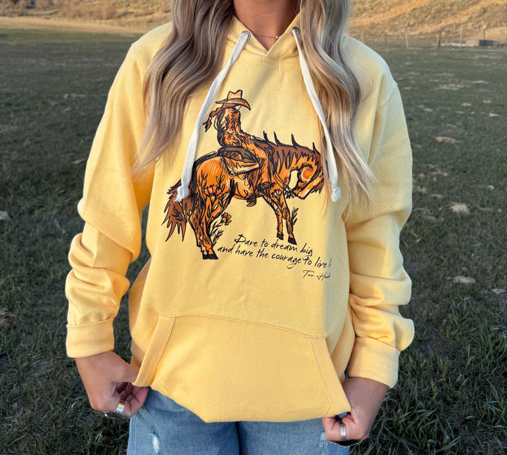 A person wearing a yellow hoodie with a graphic of a cowgirl riding a bucking horse and the text &