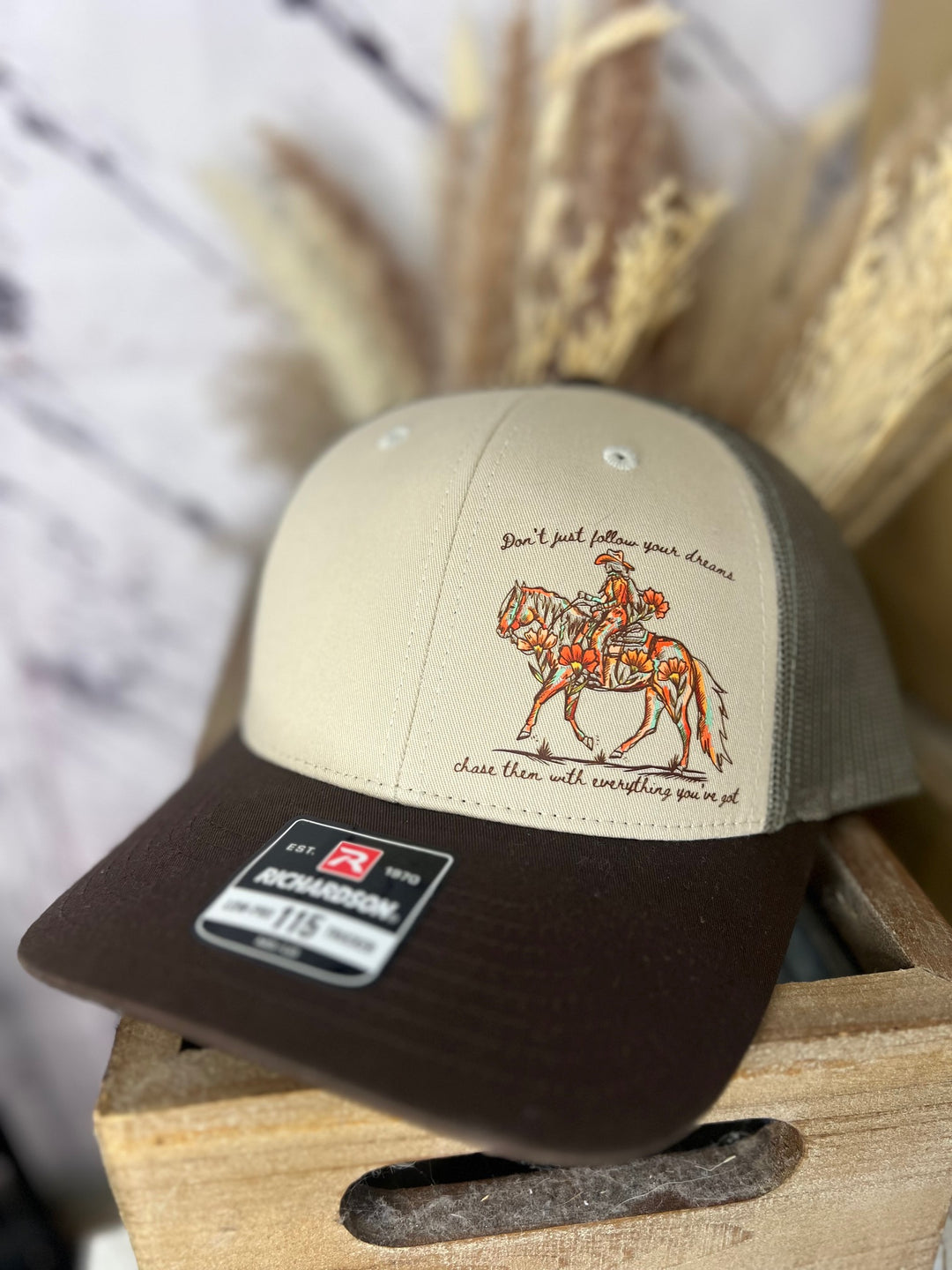 A beige and brown cowgirl ballcap with a side profile of a cowgirl riding a horse, surrounded by floral accents, and a quote that reads 'Don't just follow your dreams, chase them with everything you got.'.