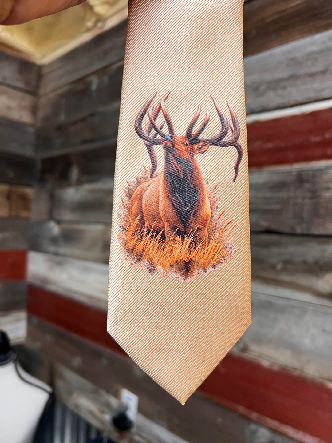 A beige textured necktie featuring a detailed graphic print of an elk deer standing in golden grass, held up against a rustic wooden wall background.