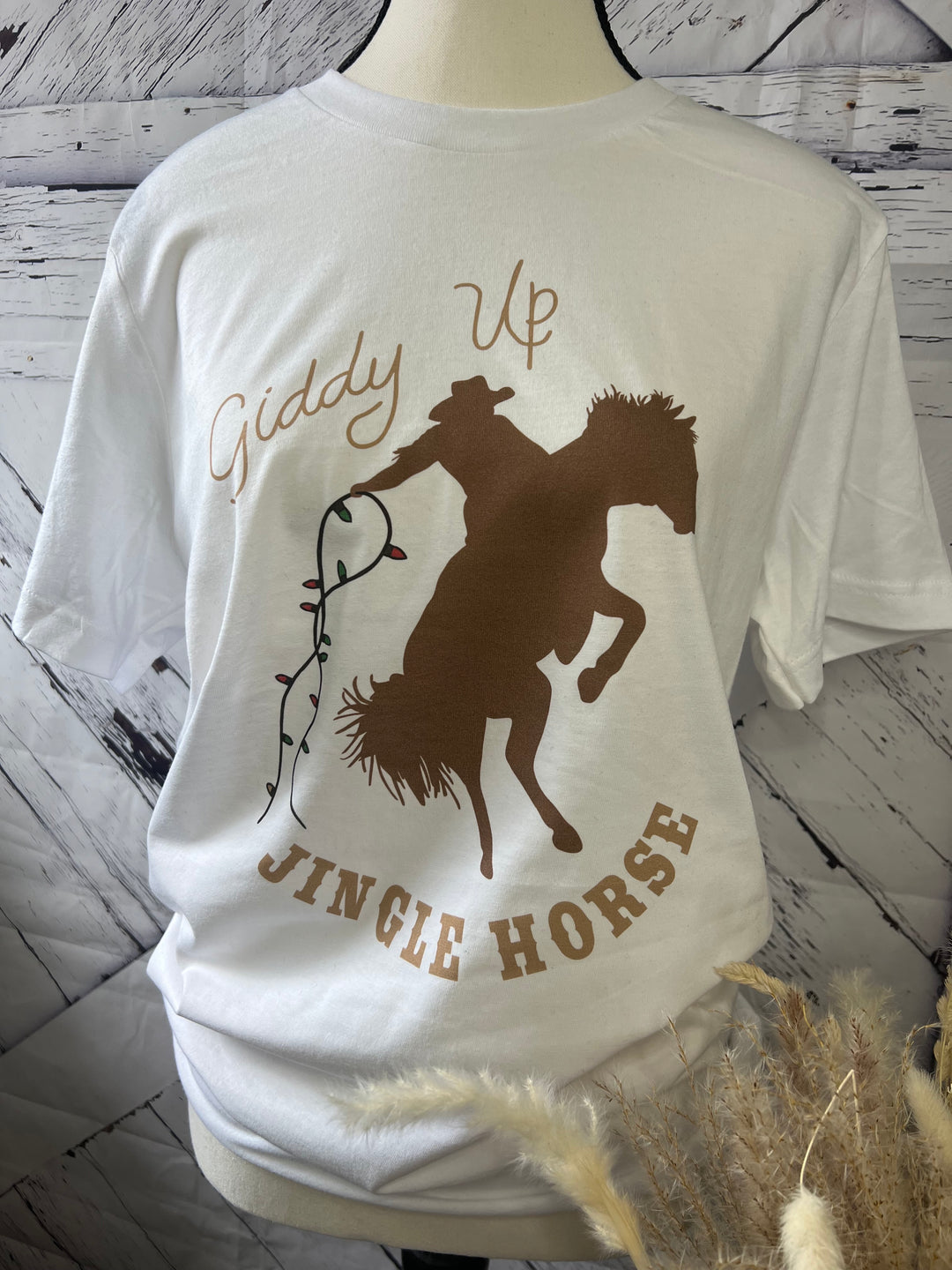 White crewneck t-shirt with 'Giddy Up' text and a graphic of a jingle horse with Christmas lights on its back, against a grey background.