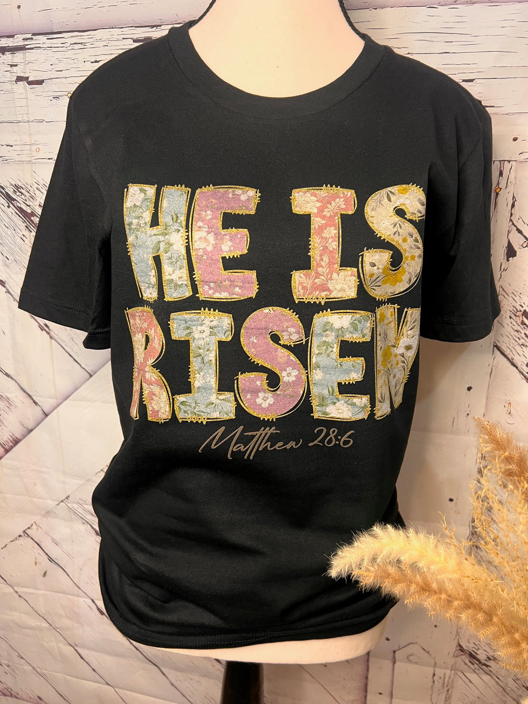 A black graphic tee with 'HE IS RISEN' written in a floral pattern with a small subtext 'Matthew 28:6' on the lower part of the tee.