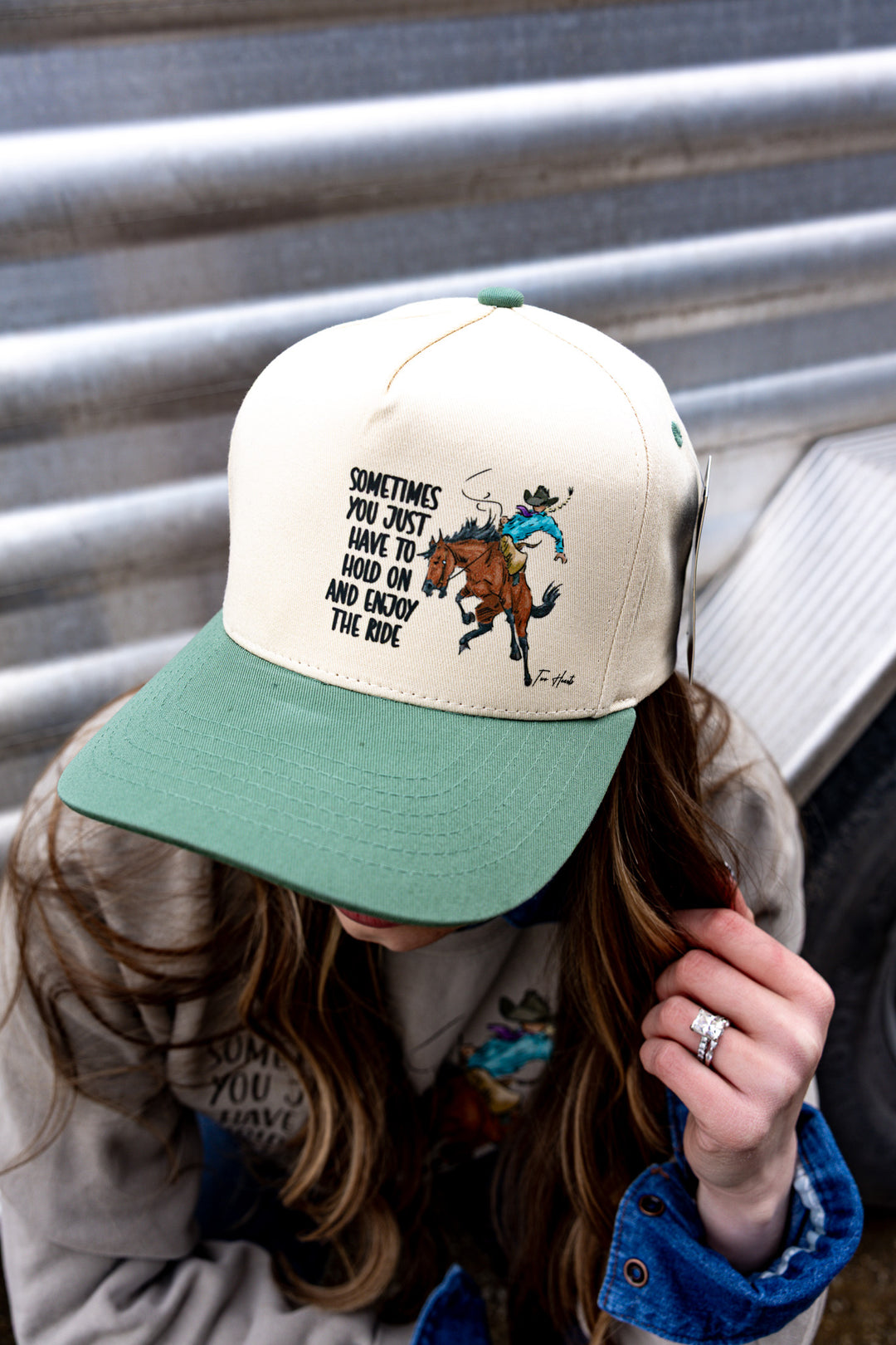 A person wearing a beige and green trucker hat with a cowgirl riding a bronc illustration on it.
