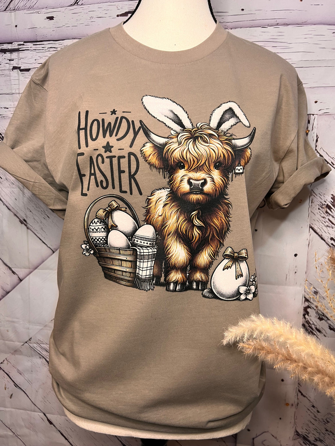 A beige t-shirt with a graphic print of a Highland cow with bunny ears and the text 'Howdy Easter'.