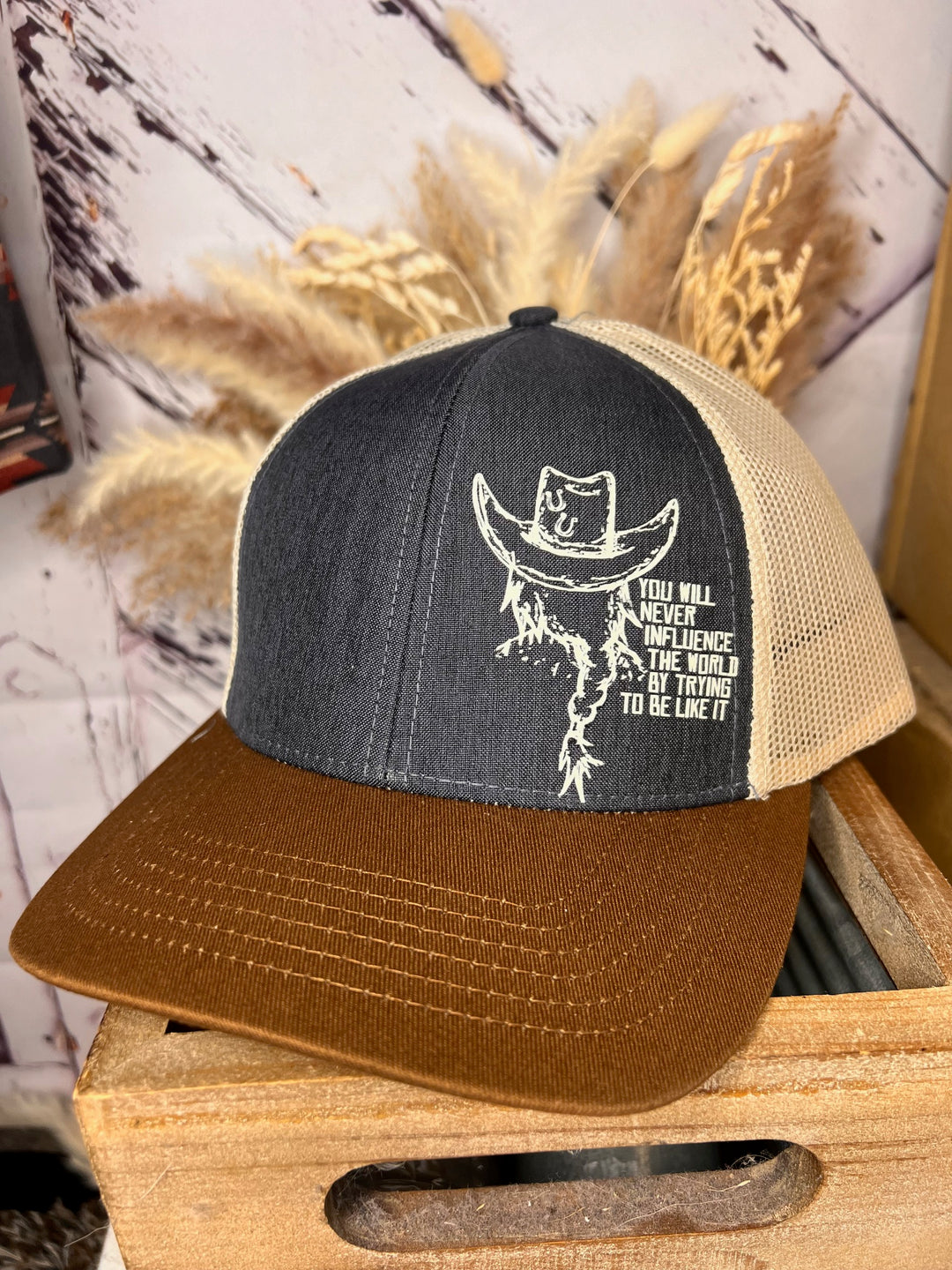 A beige, brown, and blue trucker ballcap with a cowgirl design and text on the front panel, displayed on a wooden surface with decorative elements in the background.