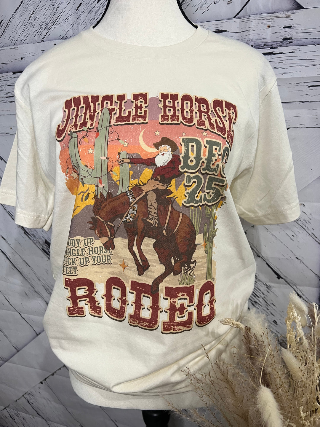 A white crew neck t-shirt with a Christmas themed graphic print featuring Santa Claus on a horse. The text 'Jingle Horse Rodeo' and the date 'DEC 25' are also visible on the shirt.