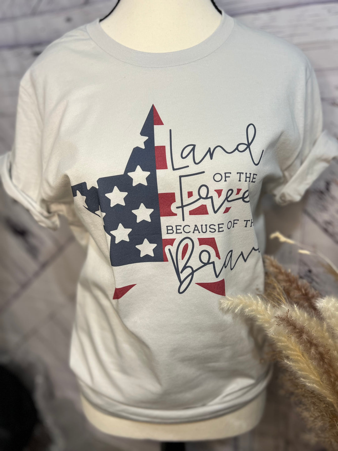 A white crew neck t-shirt with a graphic print of an American flag and the text 'LAND OF THE FREE BECAUSE OF THE BRAVE' on the front.