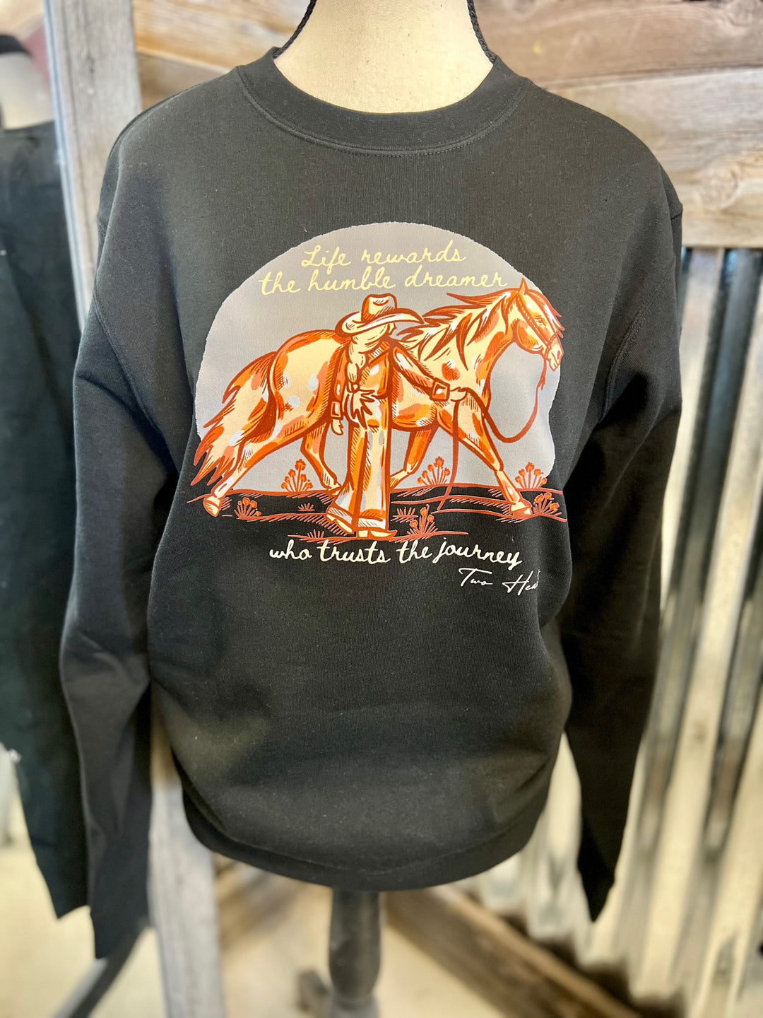 Black sweatshirt with a graphic of a person on horseback and text, displayed on a mannequin against a wooden background.