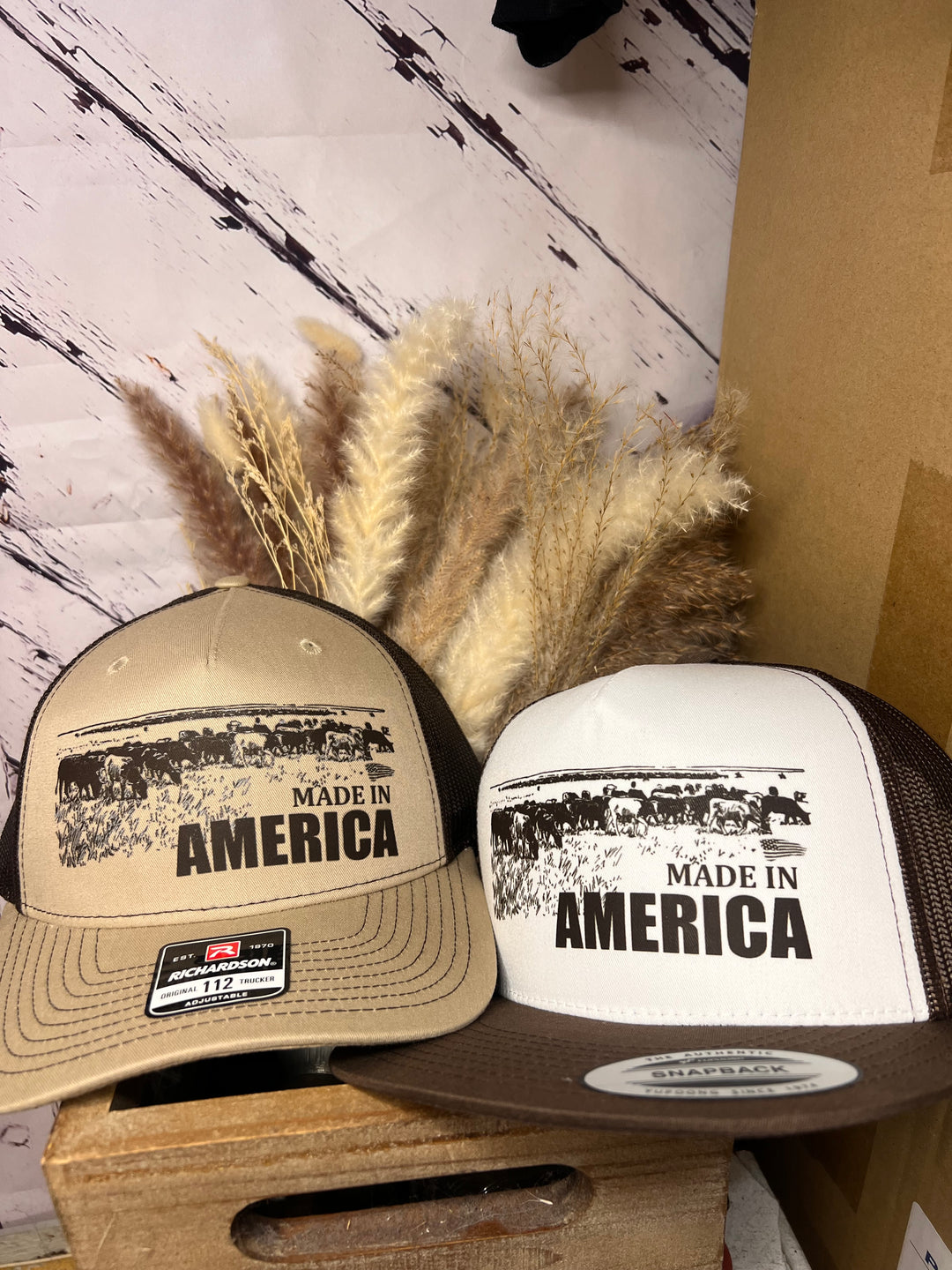 Two ballcaps, one brown and one white, with 'MADE IN AMERICA' text and an image of cowboys on horseback herding cattle.