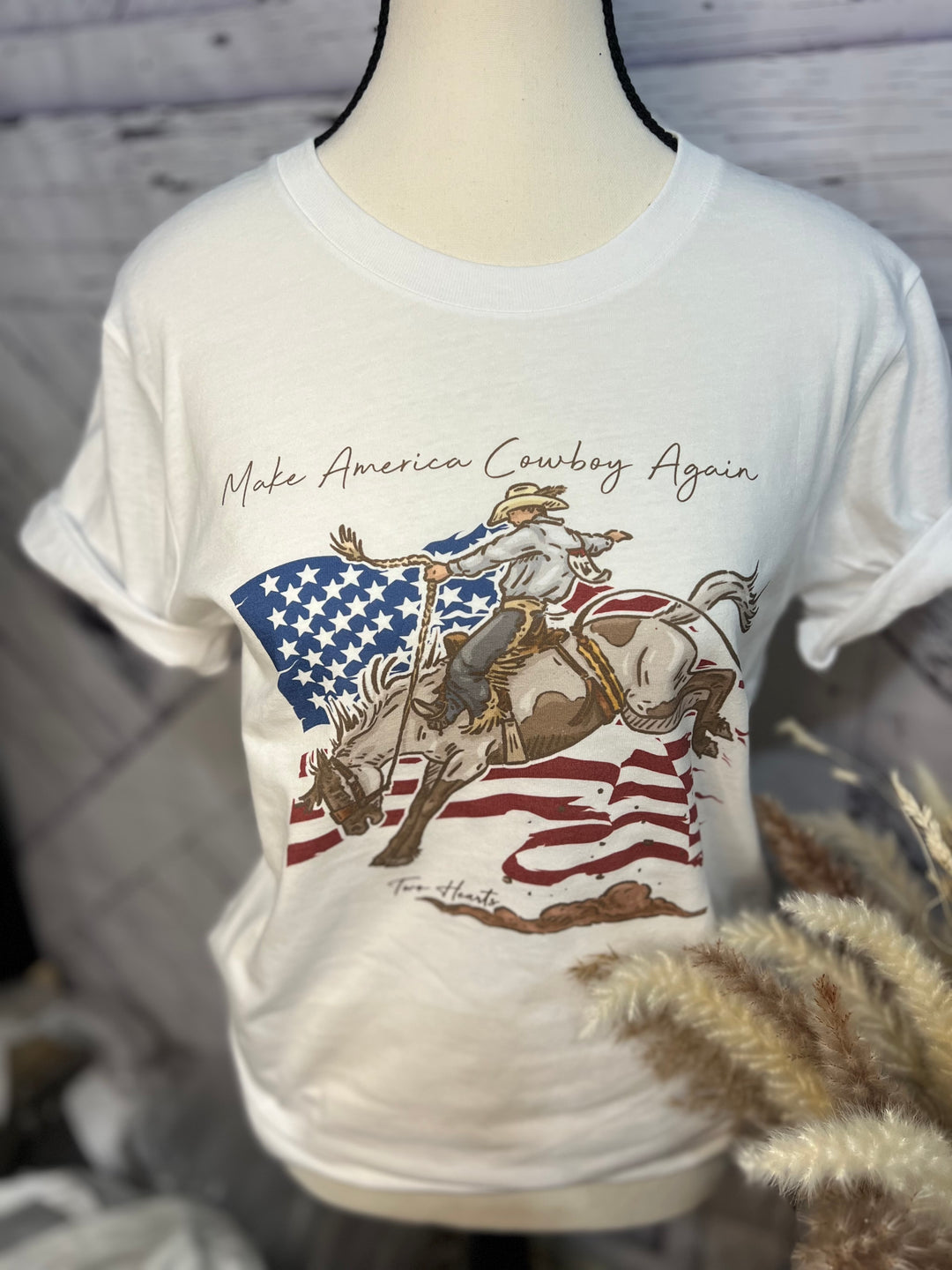 White graphic t-shirt with 'Make America Cowboy Again' text and an illustration of a cowboy bronc rider on a horse, set against a wooden background. The shirt is displayed on a mannequin, which is positioned against a backdrop featuring a patriotic-themed graphic.