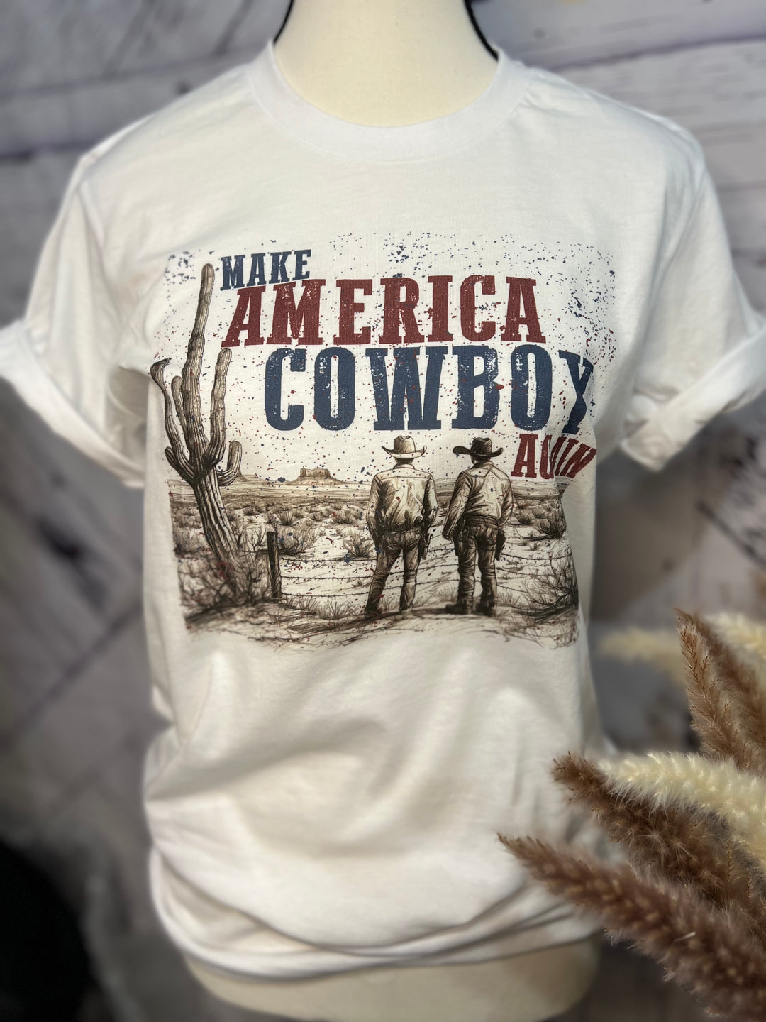 White graphic tee with 'Make America Cowboy Again' text and an illustration of cowboys in a desert landscape.