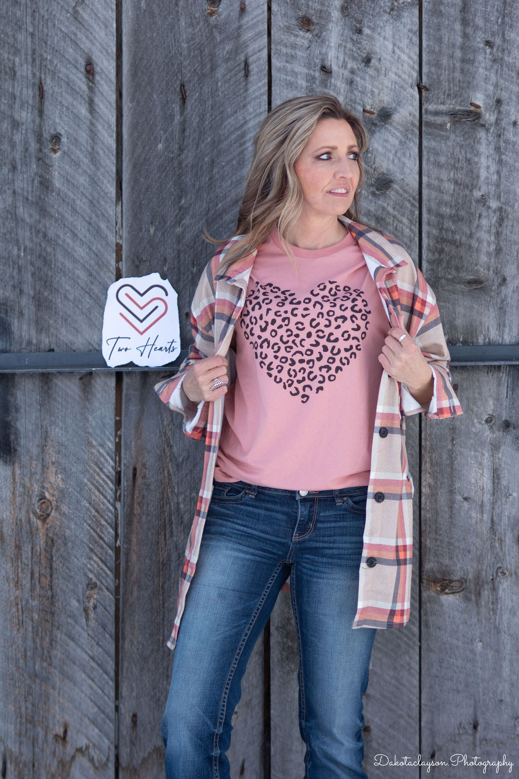 A person wearing a pink t-shirt with a cheetah heart design printed in black, paired with a plaid jacket and blue jeans, standing in front of a wooden background.