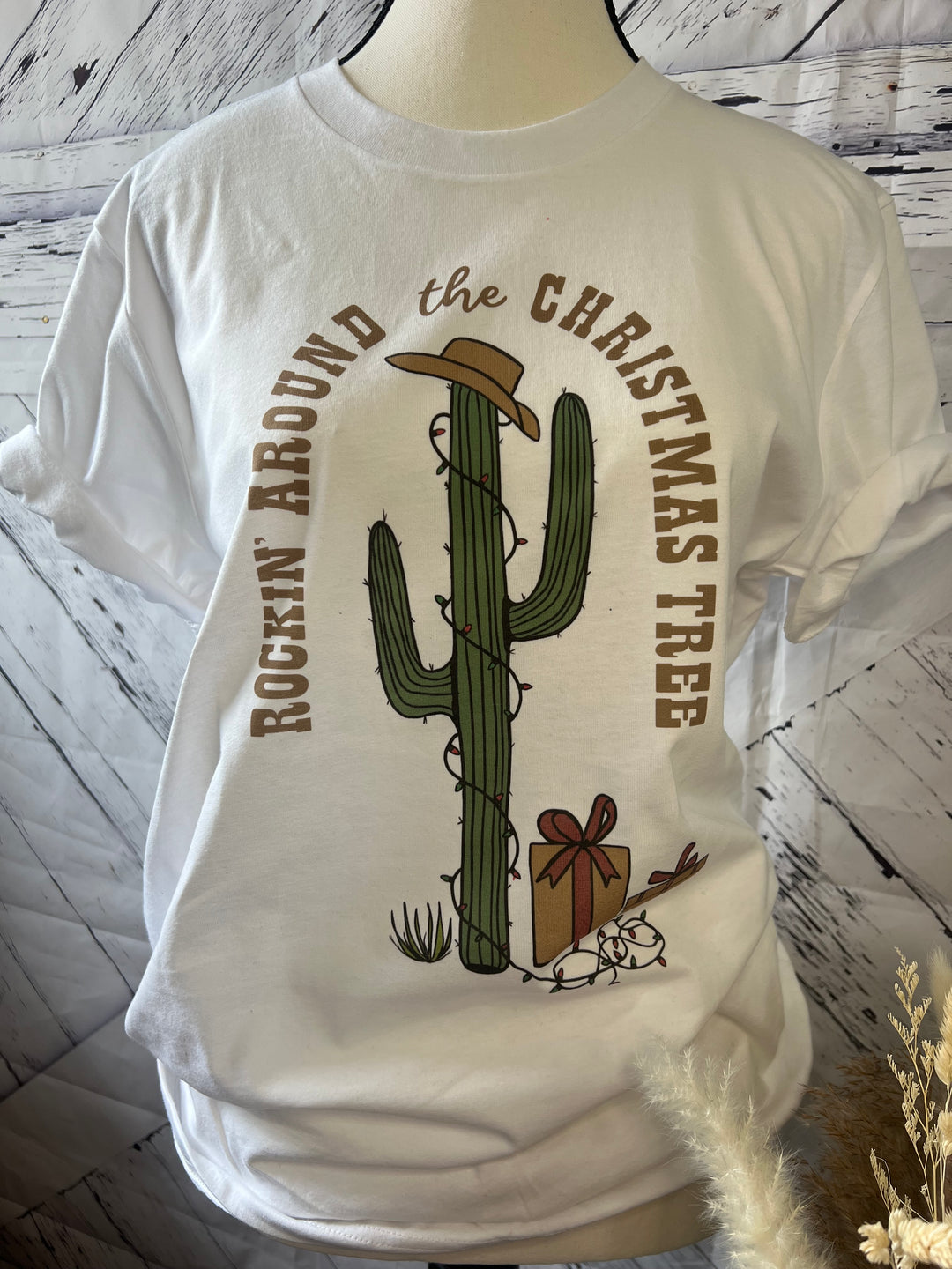 White t-shirt with 'Rockin Around Cactus' Christmas graphic design featuring a cactus in a hat and text around it.