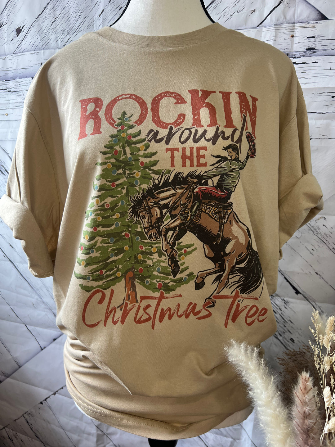 A Christmas themed graphic t-shirt featuring a man on a horse with a Christmas tree in the background, with text 'ROCKIN' AROUND THE CHRISTMAS TREE'.