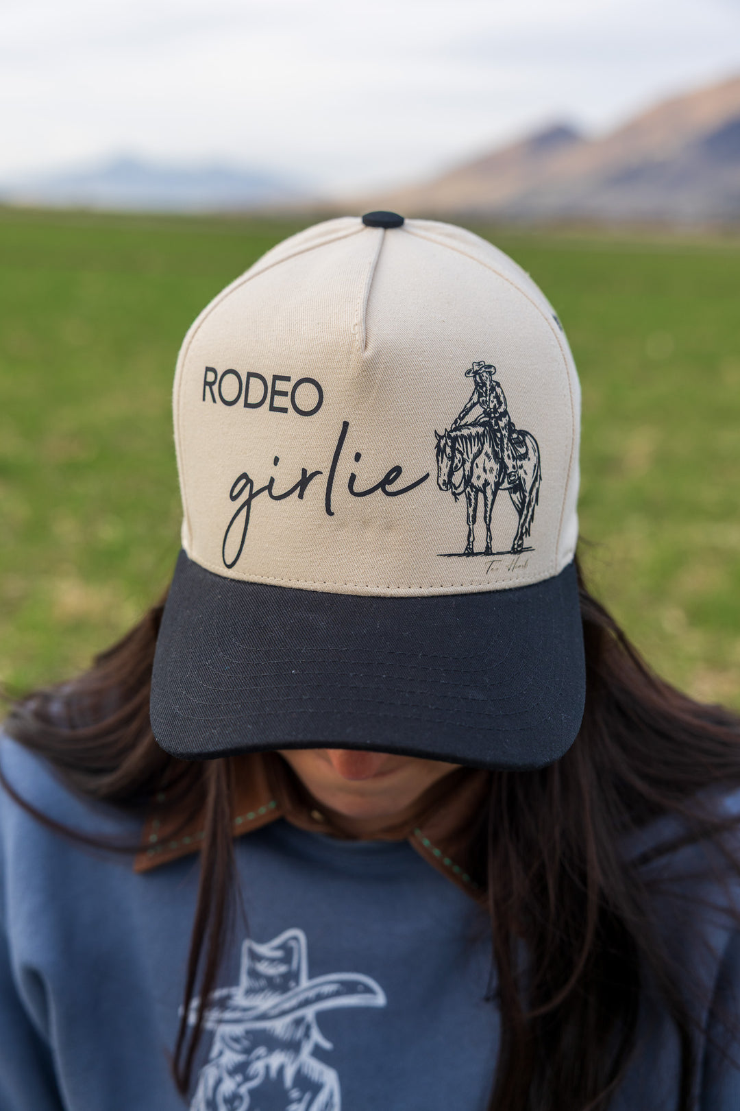 A beige and black 'Rodeo Girlie' ballcap with a graphic of a rider on a horse on the front.