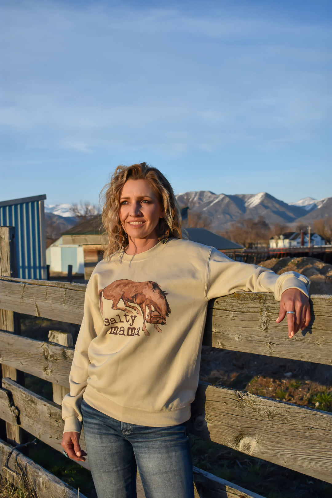 A person wearing a beige crewneck sweatshirt with a salty mama bucking horse design, standing by a wooden fence with mountains in the background.