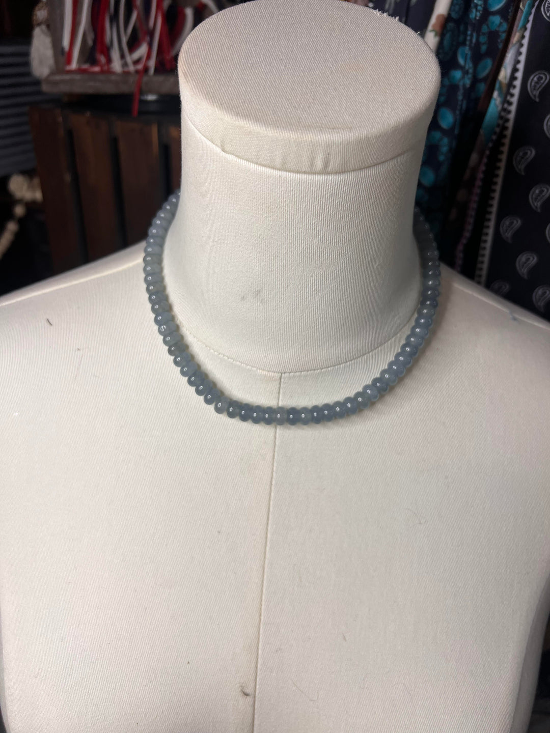 A light blue-gray beaded necklace displayed on a white fabric mannequin bust, with a blurred shop background.