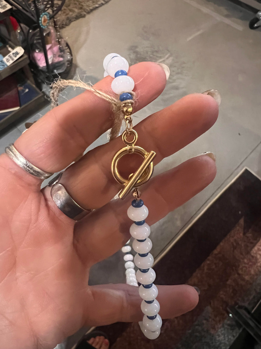A close-up of a hand holding a beaded keychain or phone strap with white and blue beads, a gold toggle clasp, and a gold ring accent, set against a blurred indoor background.