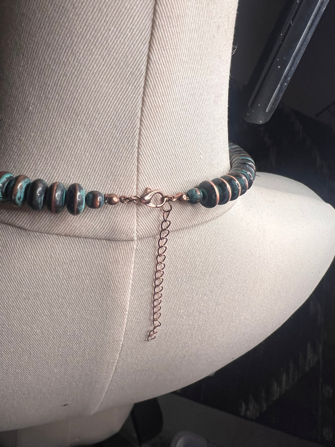A beaded bracelet with dark brown and teal-green marbled beads and a rose gold chain extender, displayed on a beige fabric-covered mannequin arm.