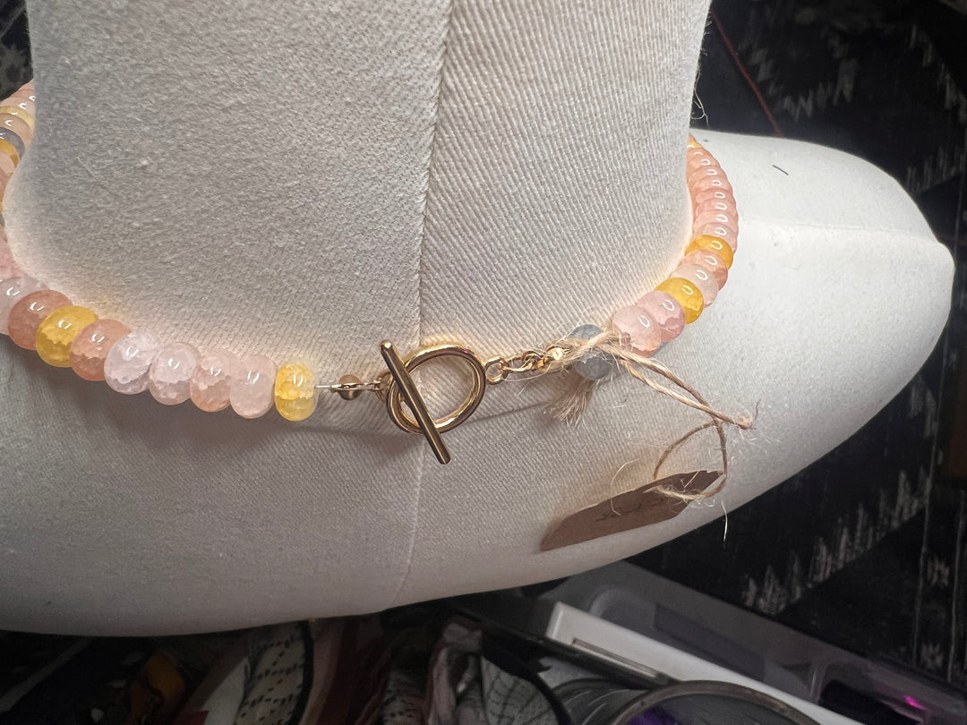 A beaded bracelet with pale pink, peach, and yellow translucent beads and a gold toggle clasp, displayed on a white fabric mannequin arm.