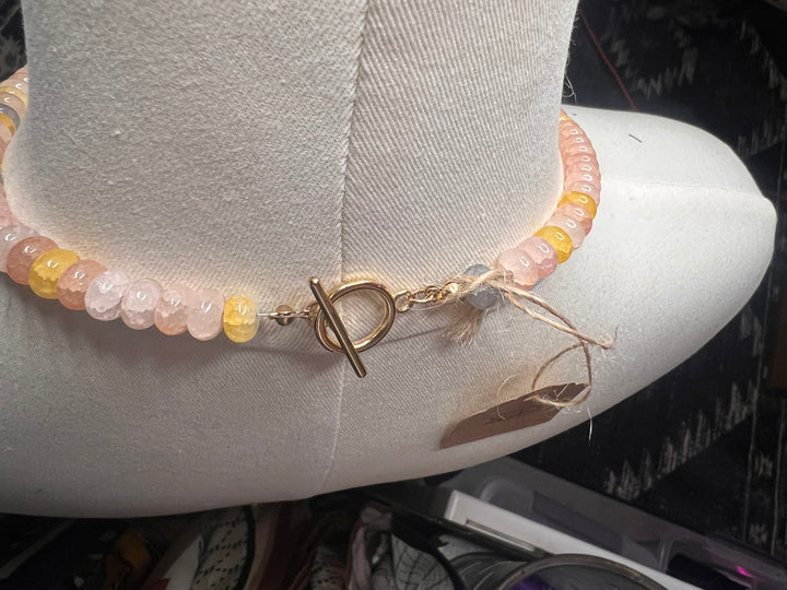 A beaded bracelet with pale pink, peach, and yellow translucent beads and a gold toggle clasp, displayed on a white fabric mannequin arm.