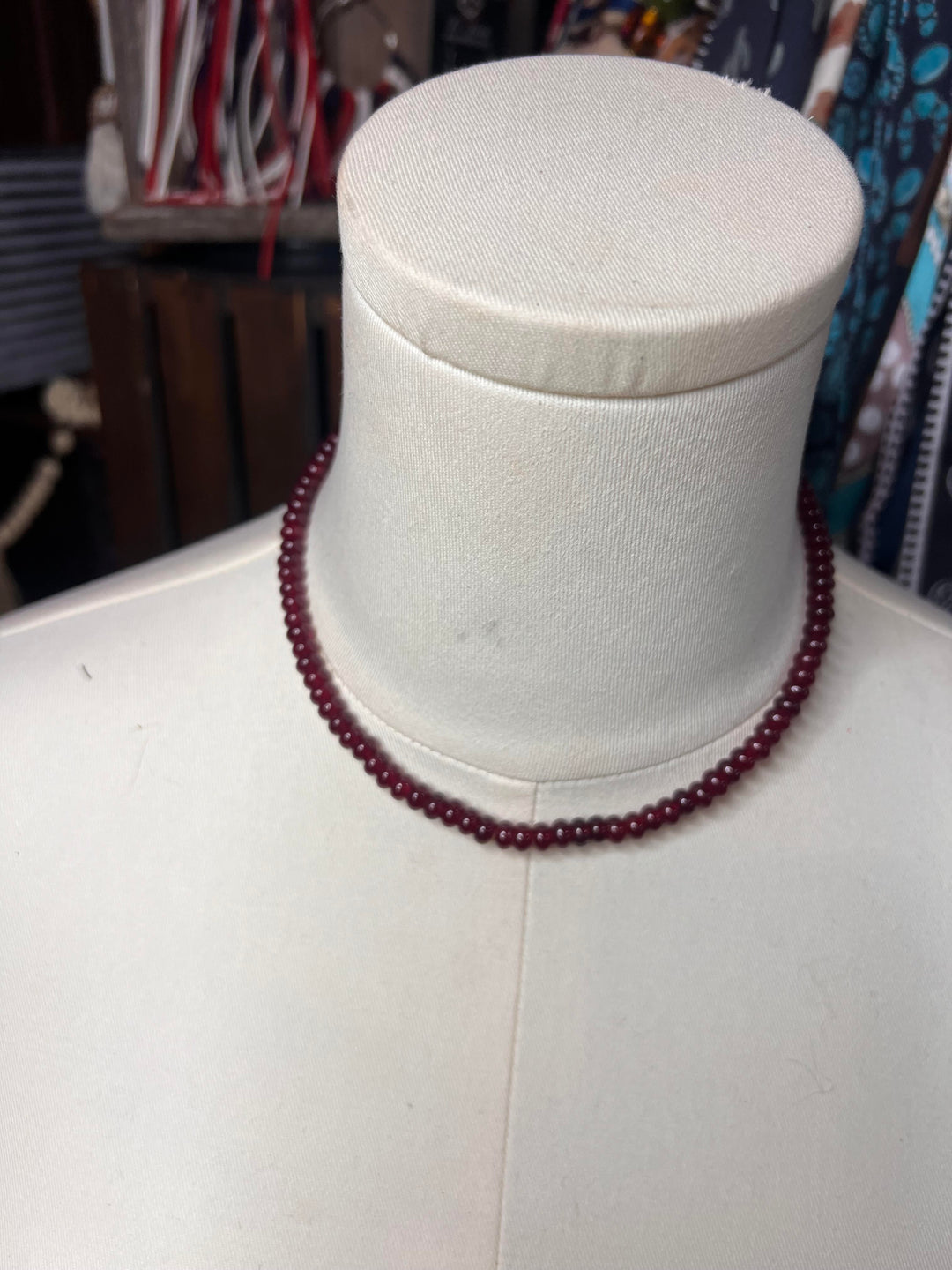 A dark red beaded necklace displayed on a cream-colored fabric mannequin bust, with a softly blurred shop background.