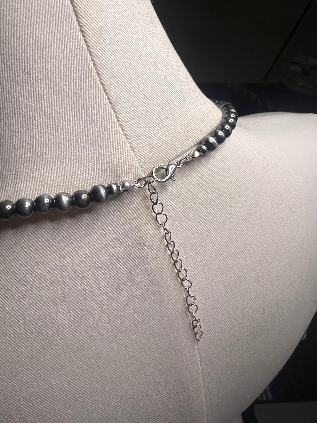 A close-up of a beaded necklace displayed on a cream fabric mannequin bust. The necklace features glossy dark gray beads and a silver-toned lobster clasp with an adjustable heart-link extension chain.