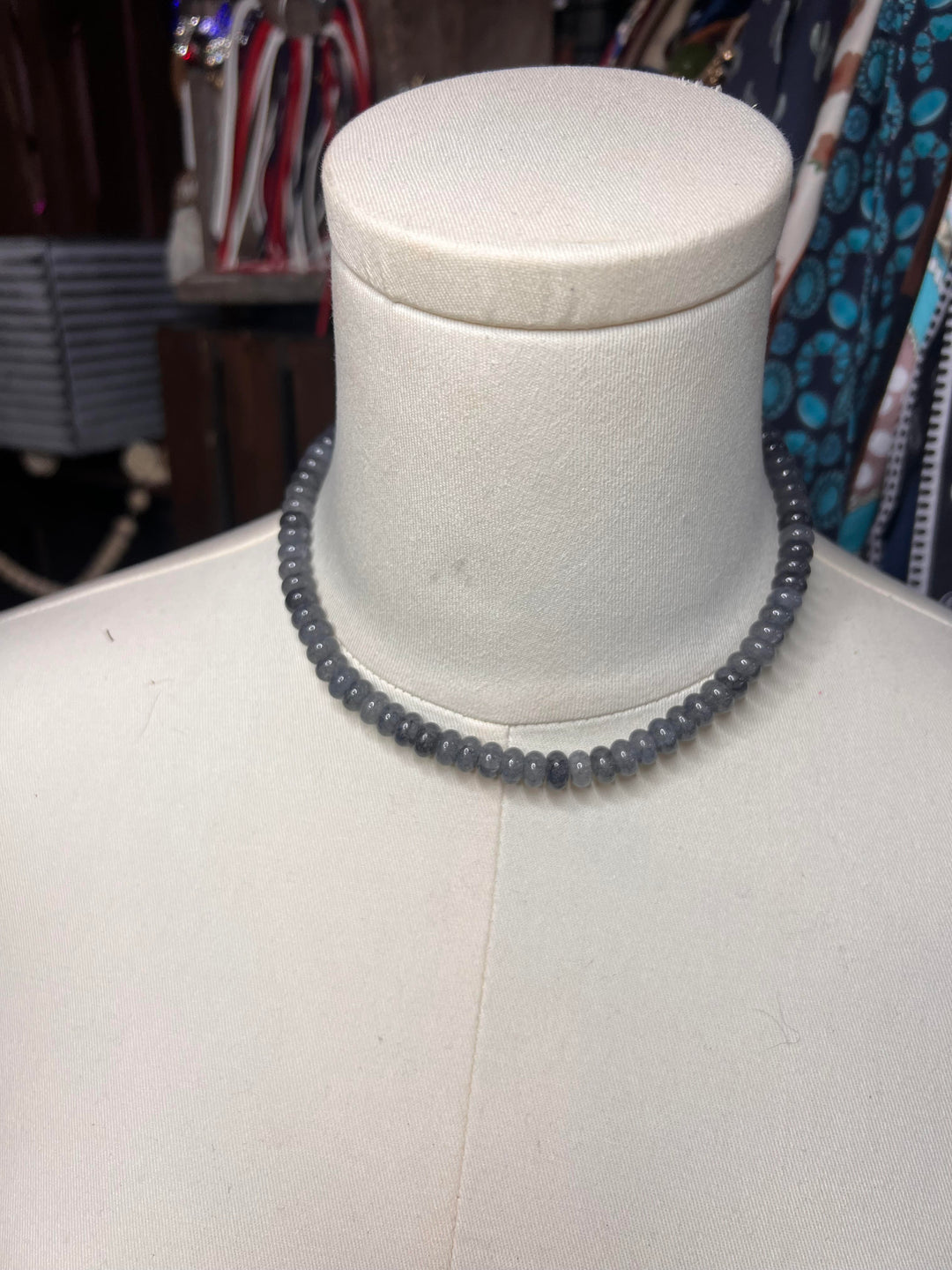 A dark gray beaded necklace displayed on a white mannequin bust, with a blurred retail shop background.