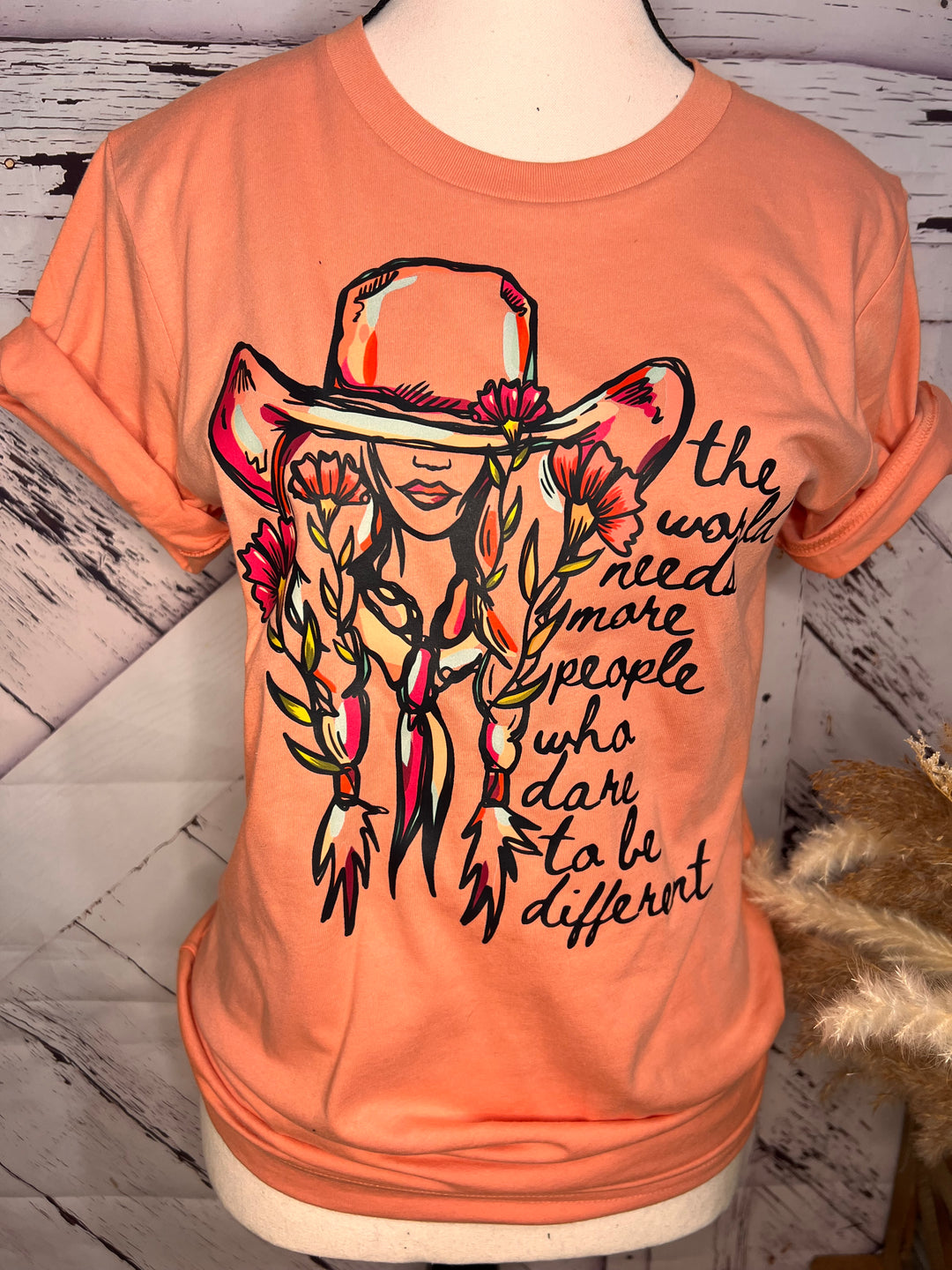 A person wearing a pink floral cowgirl tee with a quote graphic, paired with blue ripped jeans and a black jacket, sitting on a gate with a landscape in the background.