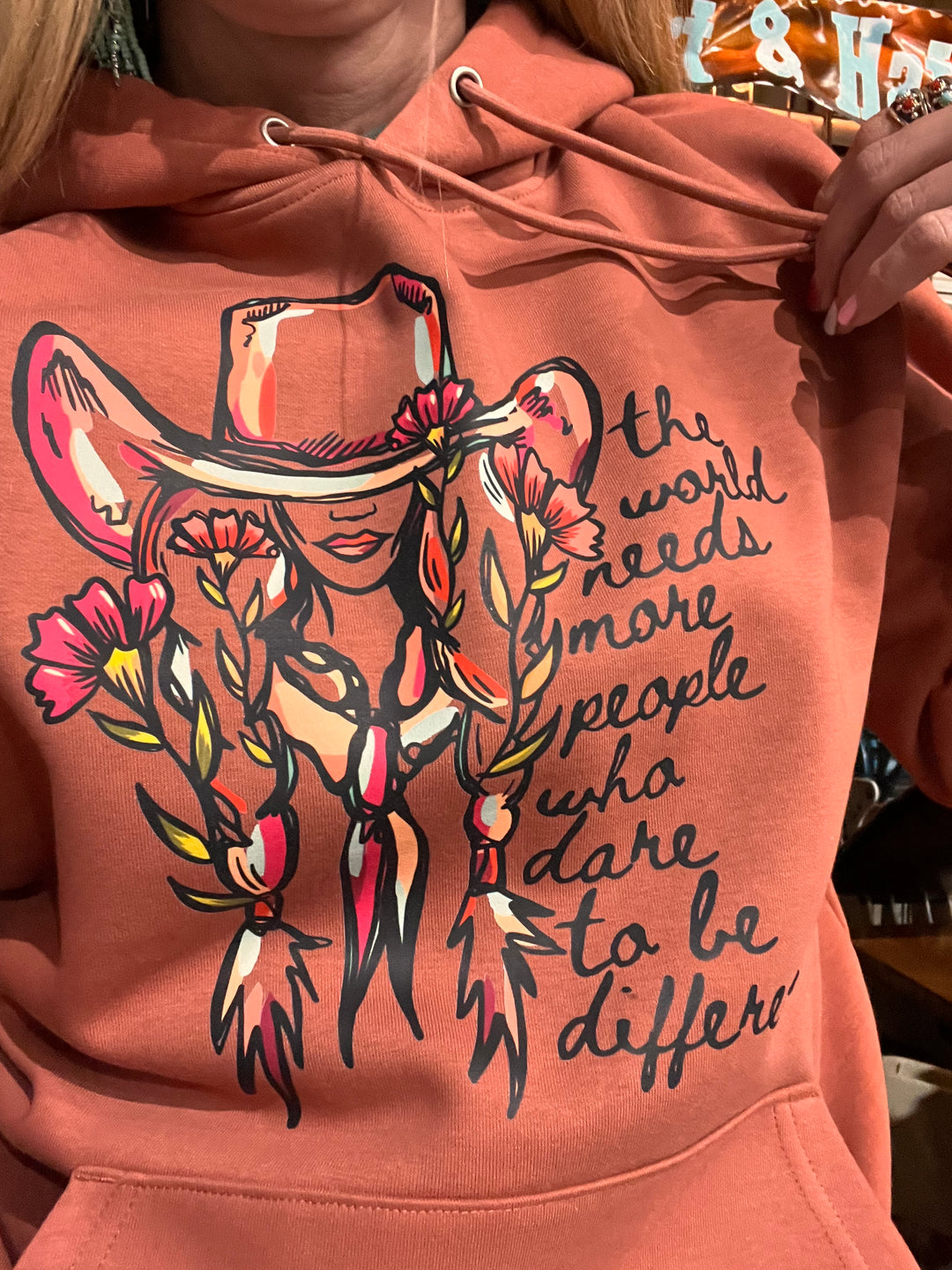 Close-up of a woman wearing a pink hoodie with a floral cowgirl design and the text 'dare to be different'.