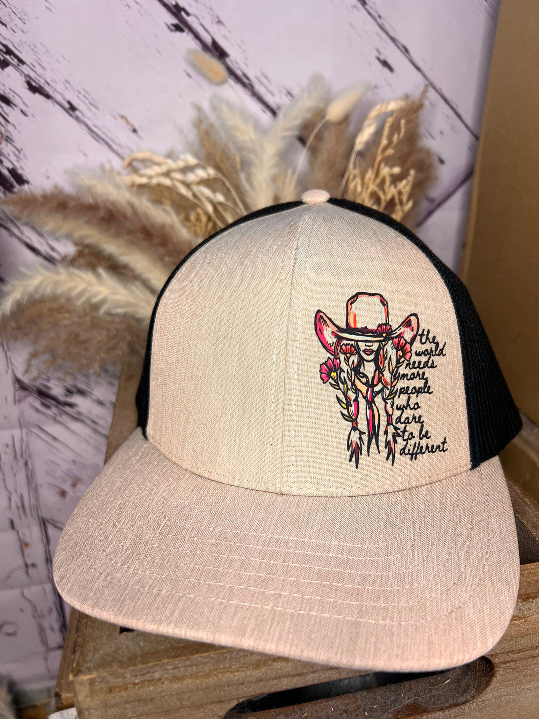 A beige and black trucker cap with a design of a cowgirl and the text 'Dare to be Different' on the front.