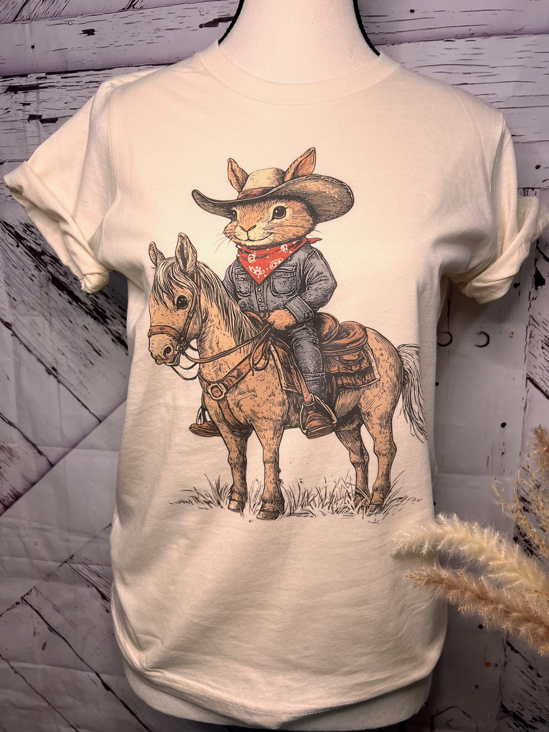 A graphic tee with a illustration of a cartoon rabbit dressed as a cowboy riding a horse, printed on a bone-colored shirt.