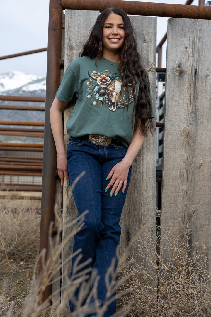 Woman wearing a green t-shirt with a graphic design, standing in front of a wooden gate with mountains in the background.