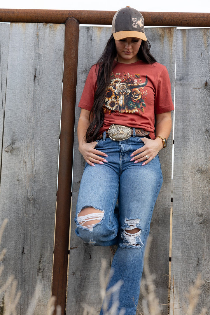 Person wearing a red graphic t-shirt, blue jeans, and a cap against a wooden fence.