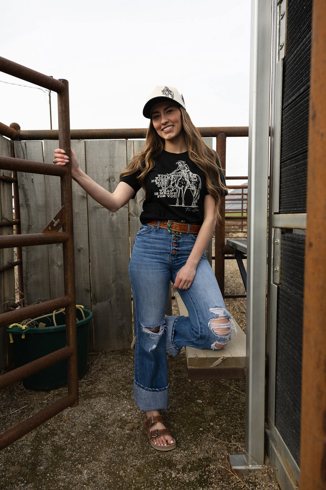 Woman standing in a barn wearing a black t-shirt with a graphic design, blue jeans, and a cap.