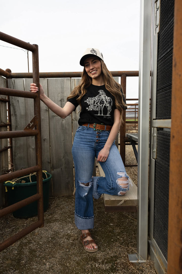 Woman standing in a barn wearing a black t-shirt with a graphic design, blue jeans, and a cap.