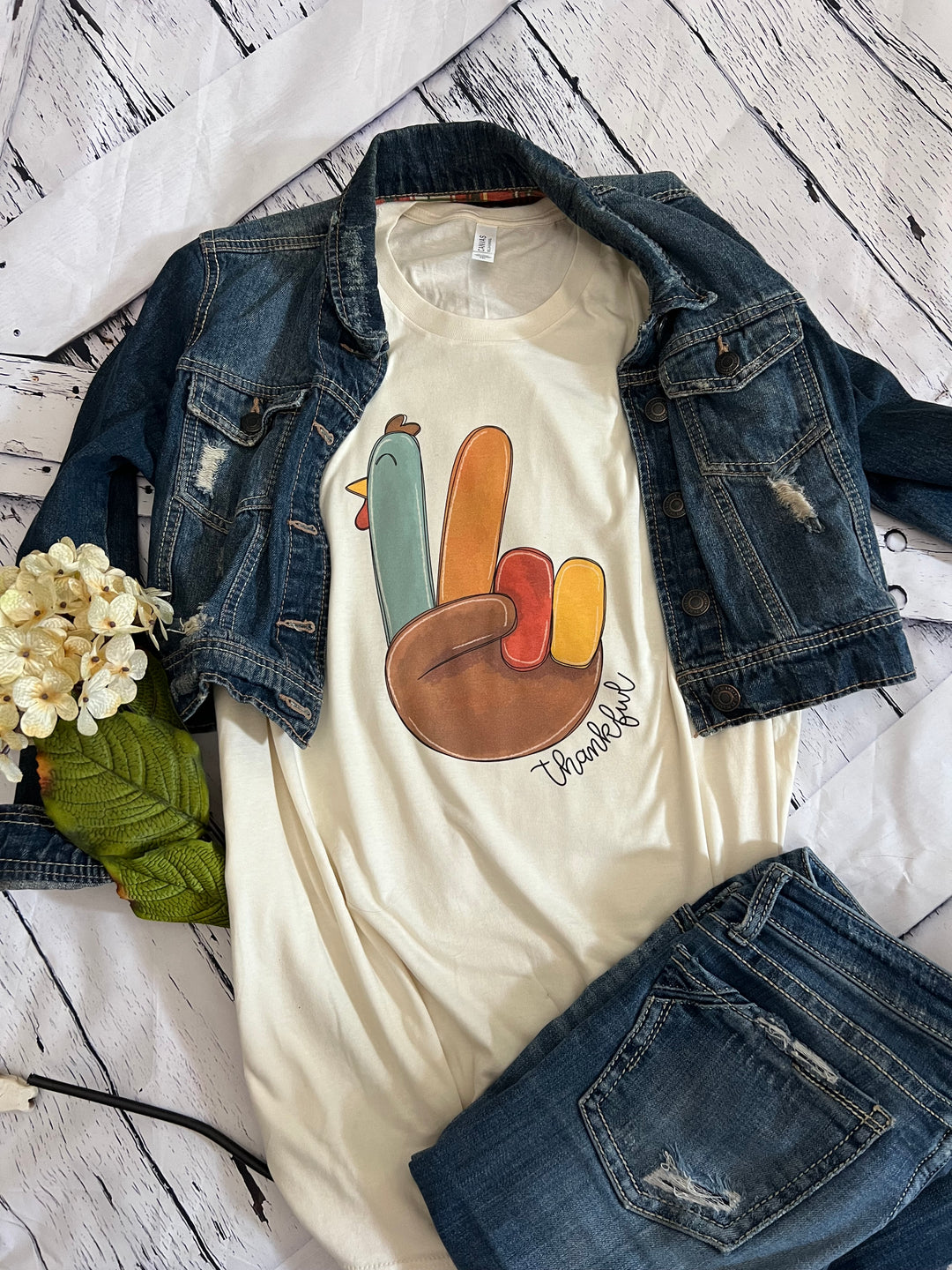 A white crewneck t-shirt with a colorful turkey design and the text 'Thankful' on the front, displayed flat with a blue denim jacket and white flowers on the side.