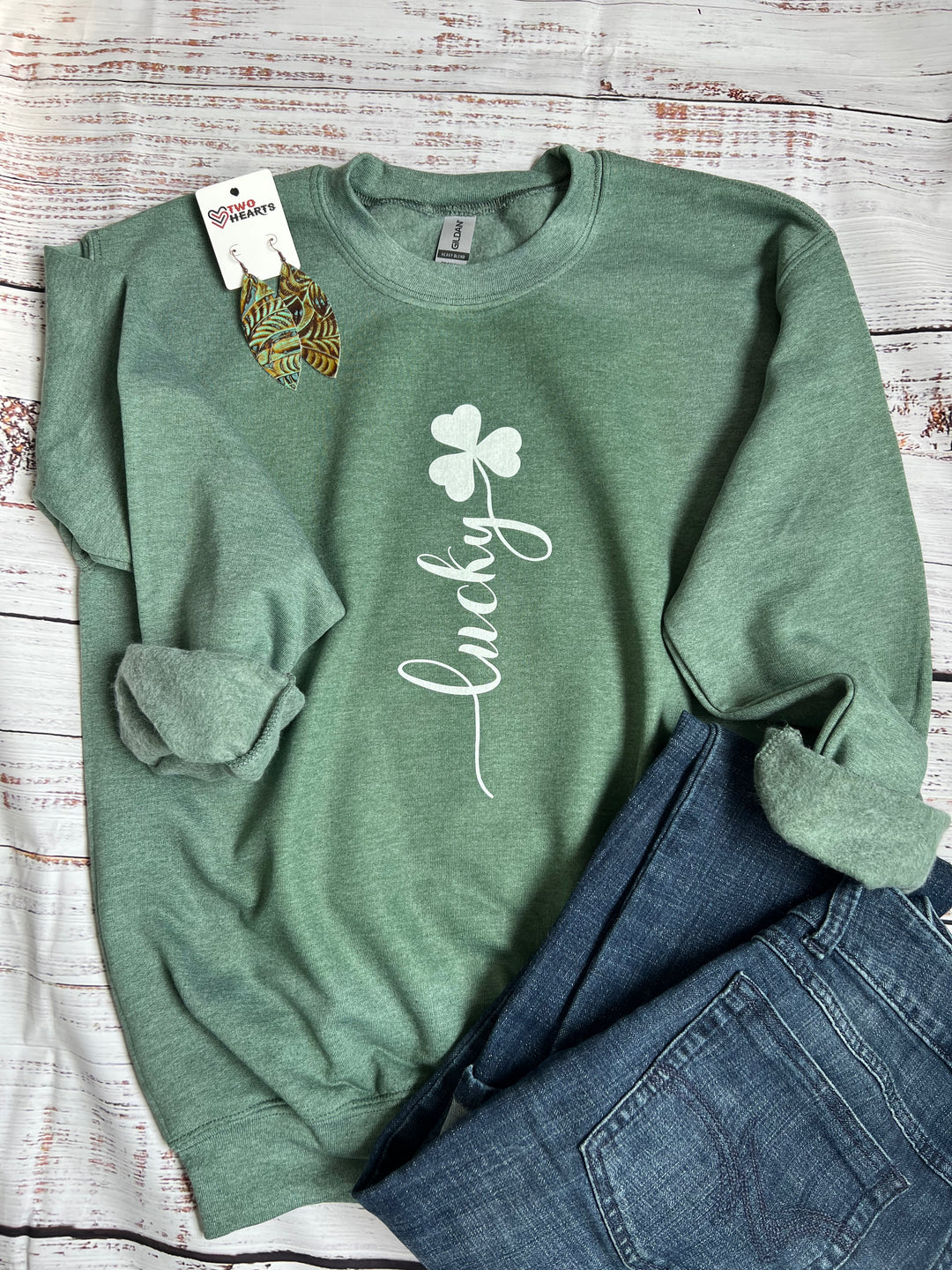 A heather green crewneck sweatshirt with the word 'lucky' written in a white script font, displayed flat with a pair of jeans.